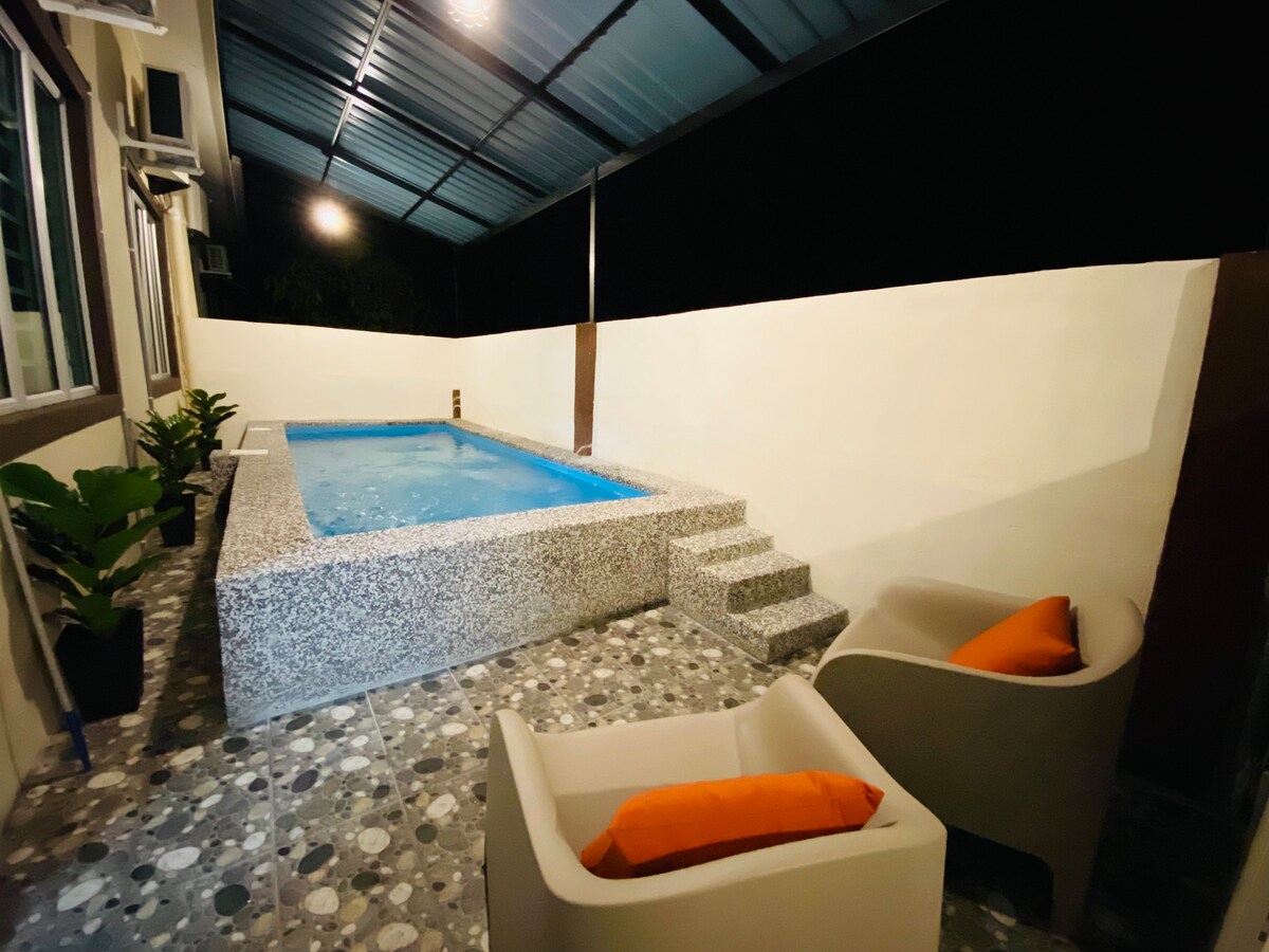 A private pool area is shown, featuring a rectangular pool surrounded by textured stone tiles. Two modern chairs with orange cushions are positioned nearby. Steps lead from the poolside to the surrounding area, which is sheltered by a metal roof.