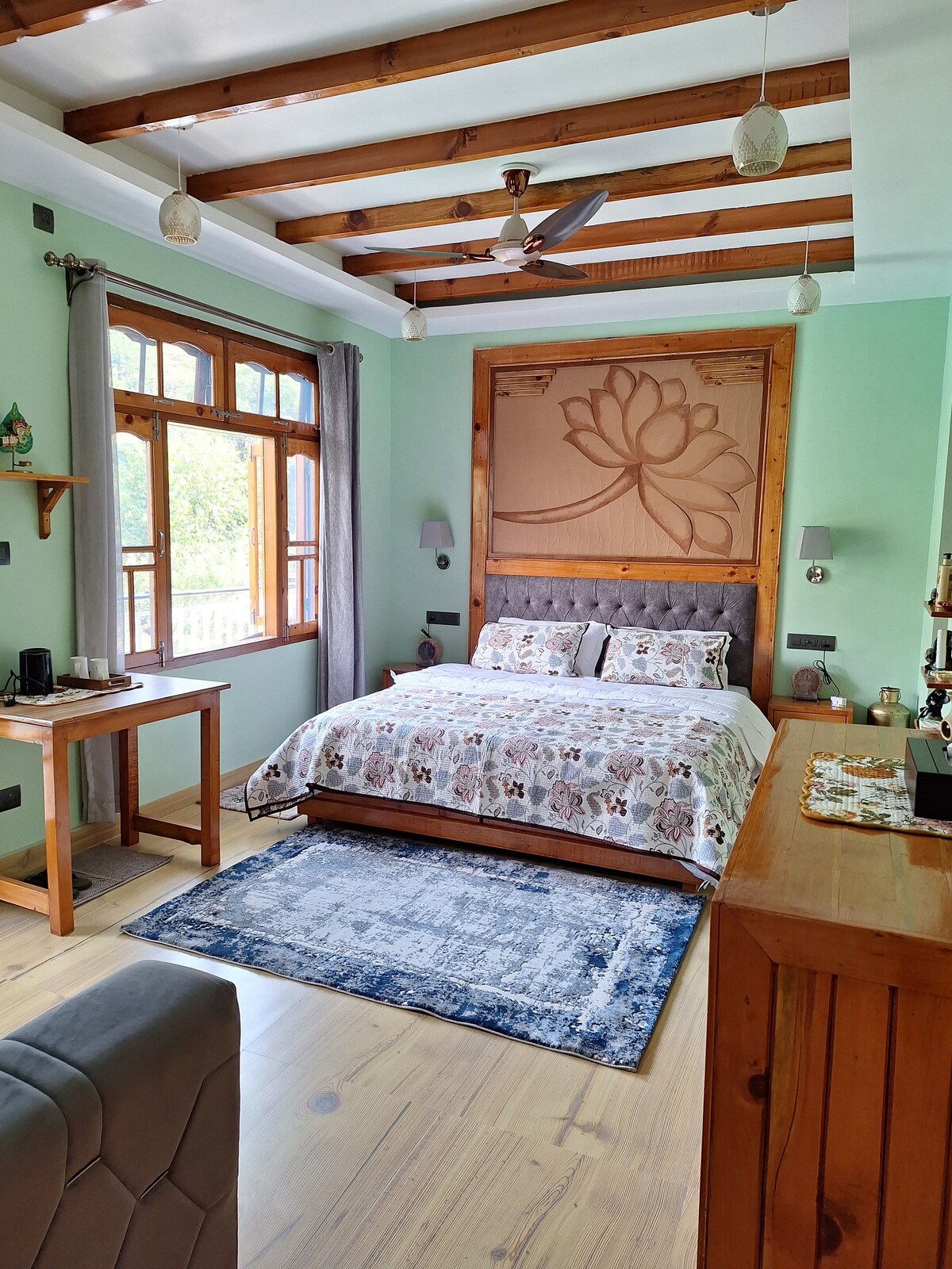 A serene room features a cozy bed with decorative bedding, complemented by a wooden headboard with a floral design. Natural light filters through large windows, and a wooden desk is positioned near the window. A plush area rug and wooden furnishings enhance the tranquil atmosphere.