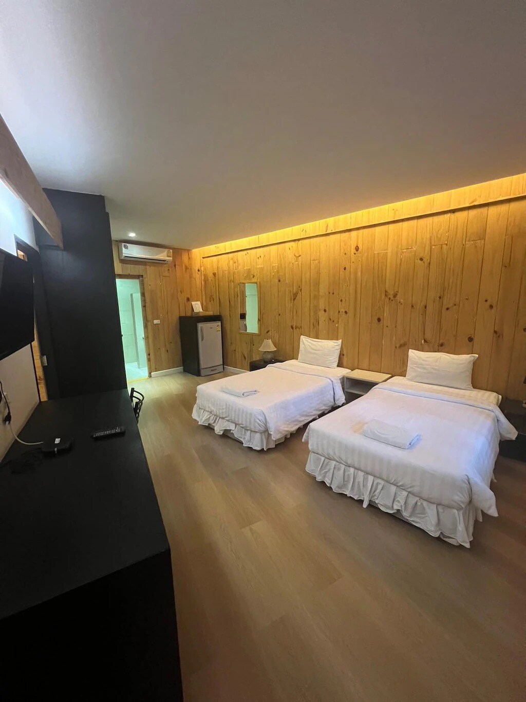 A well-lit guest room features two twin beds with white linens and decorative pillows. Wooden paneling lines the walls, contributing to a warm ambiance. A flat-screen TV is mounted opposite the beds, with a small refrigerator located in the corner.