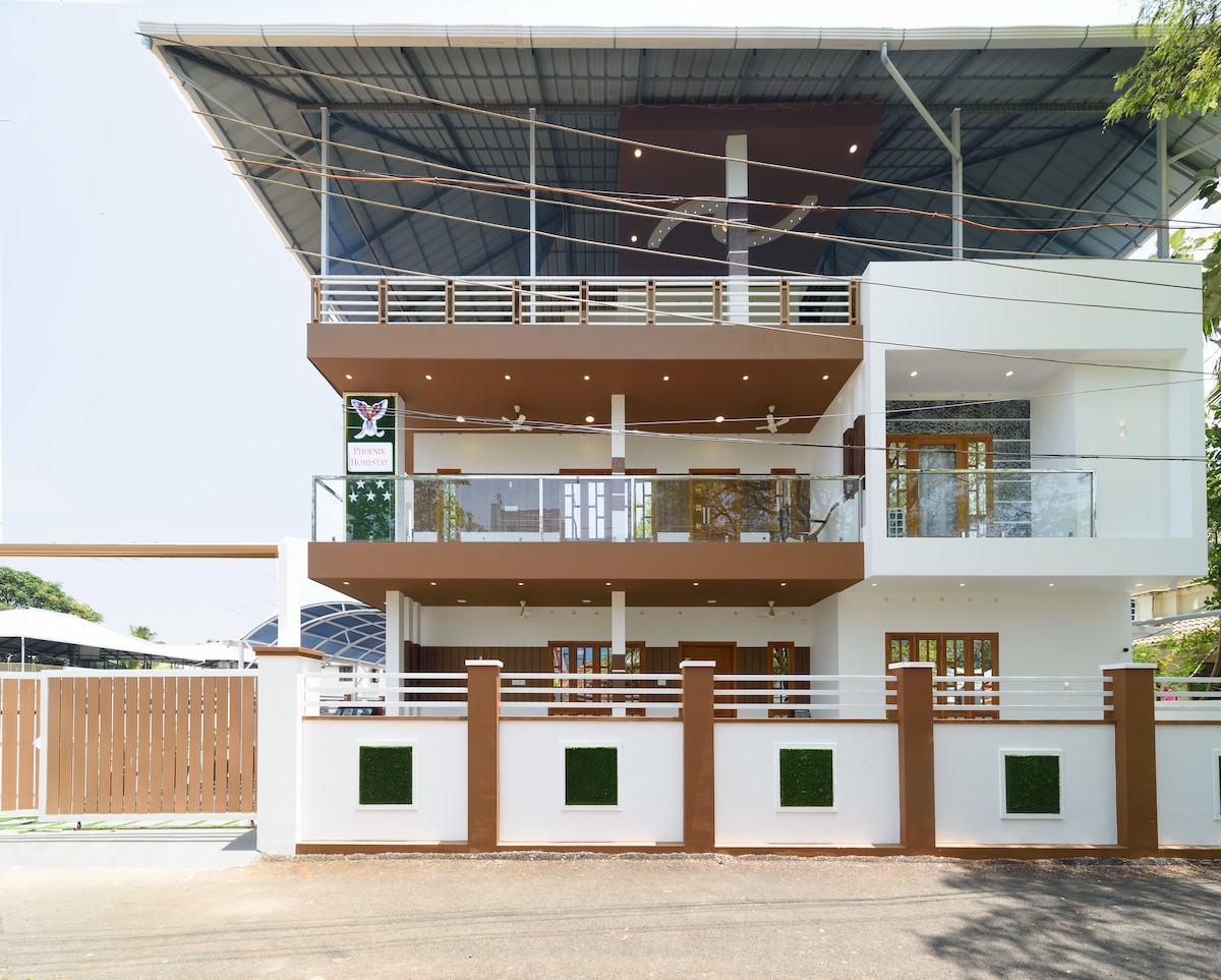 A modern three-story building is presented, highlighted by a white and brown facade. A balcony with glass railings extends from the upper levels, and a welcoming entrance is marked by decorative signage. The surrounding area is landscaped, with neatly maintained greenery.