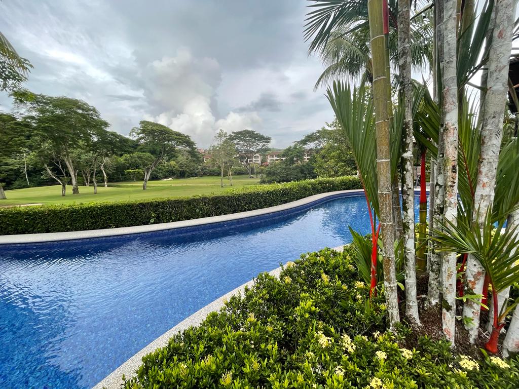 A serene swimming pool curves through manicured landscaping, featuring lush greenery and palm trees. The water reflects the overcast sky, creating a calm ambiance. The surrounding gardens enhance the natural setting, inviting relaxation.
