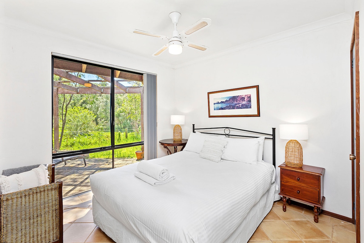 חדר שינה שליו עם מסגרת מיטה ממתכת שחורה עם מצעים לבנים חדים, שידות ליד המיטה עם מנורות קש וריצוף אריחים. כולל מאוורר תקרה וחלונות גדולים עם נוף שופע לגינה ולפאטיו.