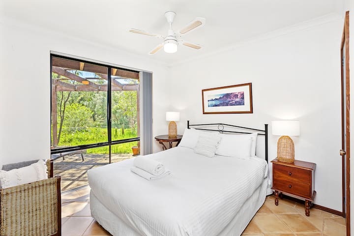 Mirna spavaća soba s crnim metalnim okvirom kreveta s svježom bijelom posteljinom, noćnim ormarićima s pletenim svjetiljkama i popločanim podovima. Uključuje stropni ventilator i velike prozore s bujnim pogledom na vrt i terasu.