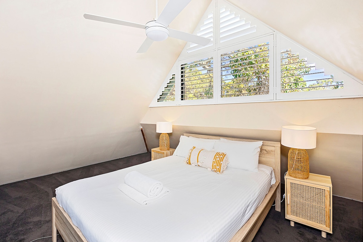 חדר שינה נעים בסגנון לופט עם תקרה דקה עם תריסי מטע, מסגרת מיטה מעץ ומצעים רכים. כולל שידות ליד המיטה עם מנורות, מאוורר תקרה וחלונות גדולים לאור טבעי.