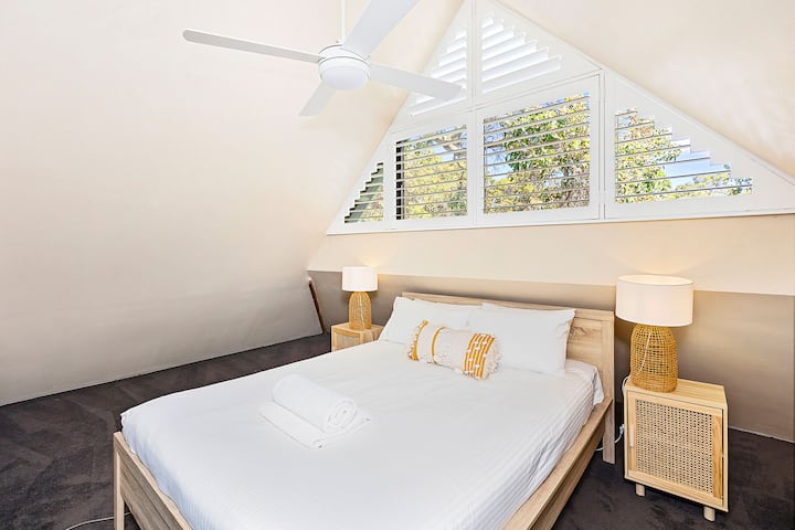 Udobna spavaća soba u potkrovlju sa stropom i roletama na plantažama, drvenim okvirom kreveta i mekom posteljinom. Uključuje noćne ormariće sa svjetiljkama, stropni ventilator i velike prozore za prirodno svjetlo.