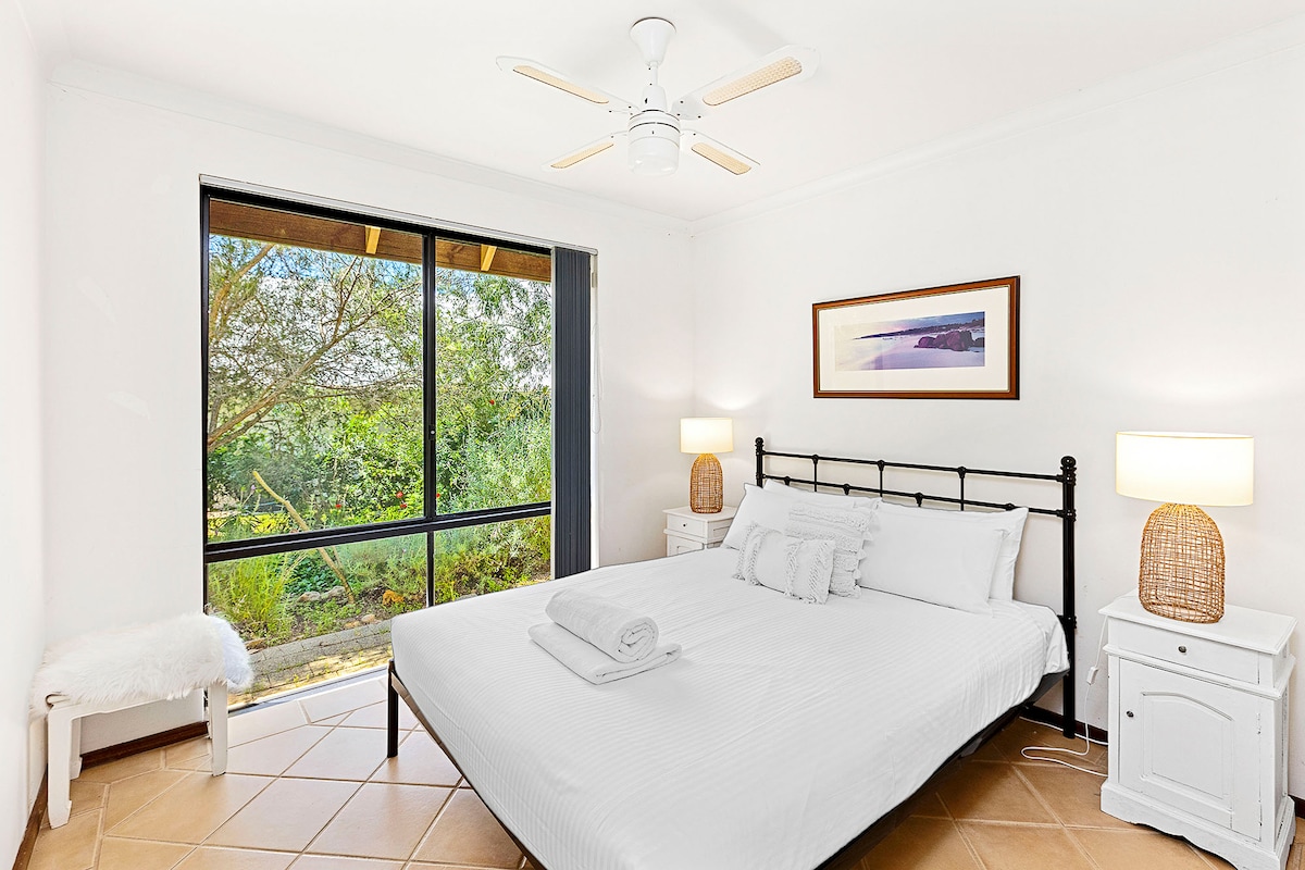 חדר שינה מואר עם מסגרת מיטה ממתכת שחורה עם מצעים לבנים חדים, שידות ליד המיטה עם מנורות קש וריצוף אריחים. כולל מאוורר תקרה וחלון גדול עם נוף שליו לגינה.