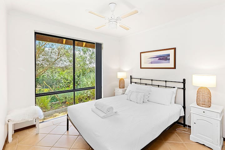 Svijetla spavaća soba s crnim metalnim okvirom kreveta s svježom bijelom posteljinom, noćnim ormarićima s pletenim svjetiljkama i popločanim podovima. Uključuje stropni ventilator i veliki prozor s mirnim pogledom na vrt.