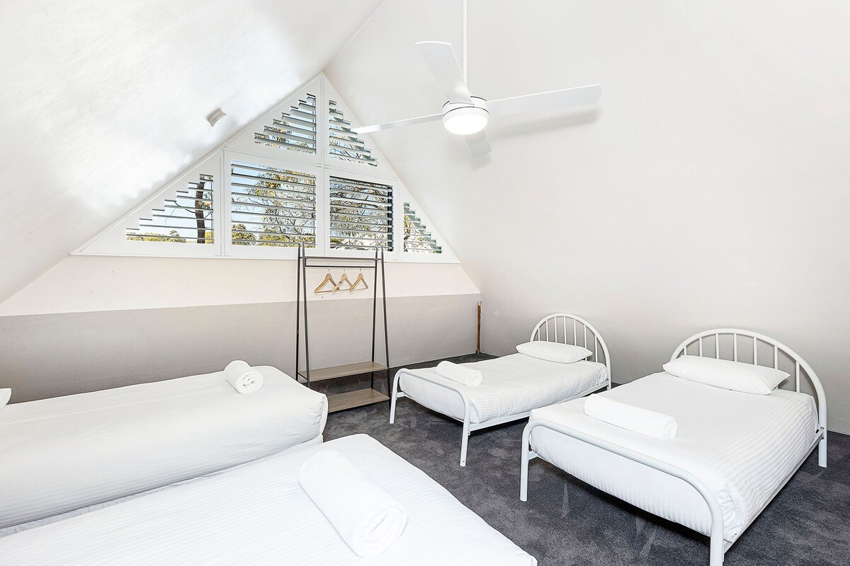 חדר שינה מרווח בסגנון לופט עם ארבע מיטות יחיד, מושלם לשהיות קבוצתיות. המאפיינים כוללים תקרות קמורות, תריסי מטע, מאוורר תקרה, שטיחים רכים וארון בגדים מינימליסטי עם קולבים.