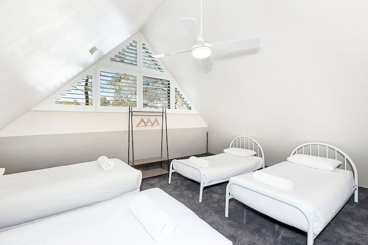 Prostrana spavaća soba u potkrovlju s četiri kreveta za jednu osobu savršena za grupne boravke. Značajke uključuju nadsvođene stropove, rolete na plantažama, stropni ventilator, mekane tepihe i minimalistički ormar s vješalicama.