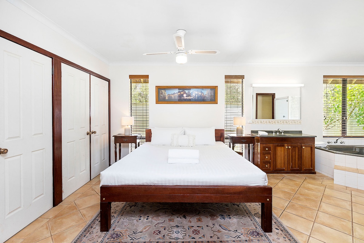 חדר שינה מרווח עם מסגרת מיטה מעץ, סוויטה עם אמבטיית ספא מובנית וכיורים כפולים. כולל ארונות גדולים, שידות ליד המיטה עם מנורות, רצפות אריחים ואור טבעי מחלונות מרובים.