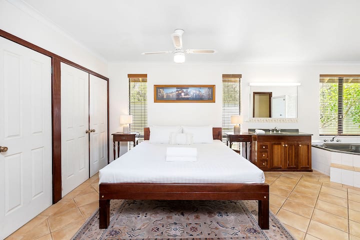 Prostrana spavaća soba s drvenim okvirom kreveta, kupaonicom s ugrađenom hidromasažnom kadom i dva umivaonika. Uključuje velike ormare, noćne ormariće sa svjetiljkama, popločane podove i prirodno svjetlo s više prozora.