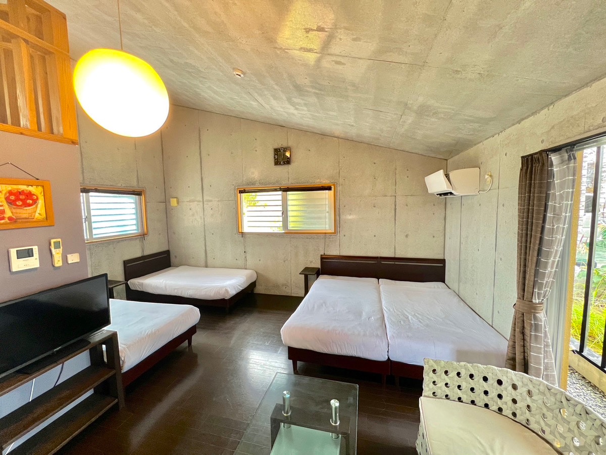 A spacious room features two large beds positioned against the wall, with a table and chairs nearby. Natural light enters through multiple windows, enhancing the neutral concrete walls and dark flooring. A television is placed on a stand, and a light fixture hangs from the ceiling.