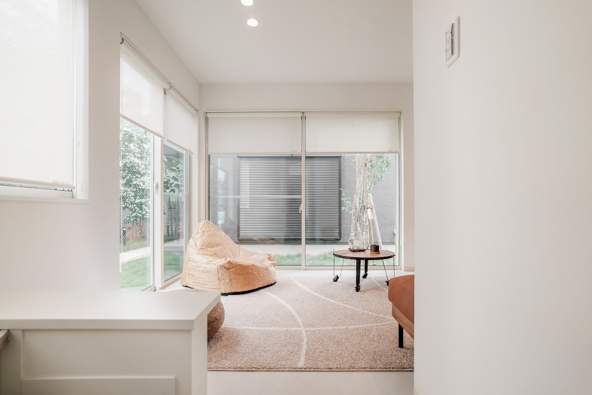 A bright and airy living space features large windows that allow natural light to fill the room. A light-colored rug covers the floor, complemented by a cozy seating area. A small round table and a relaxed chair add to the inviting atmosphere.