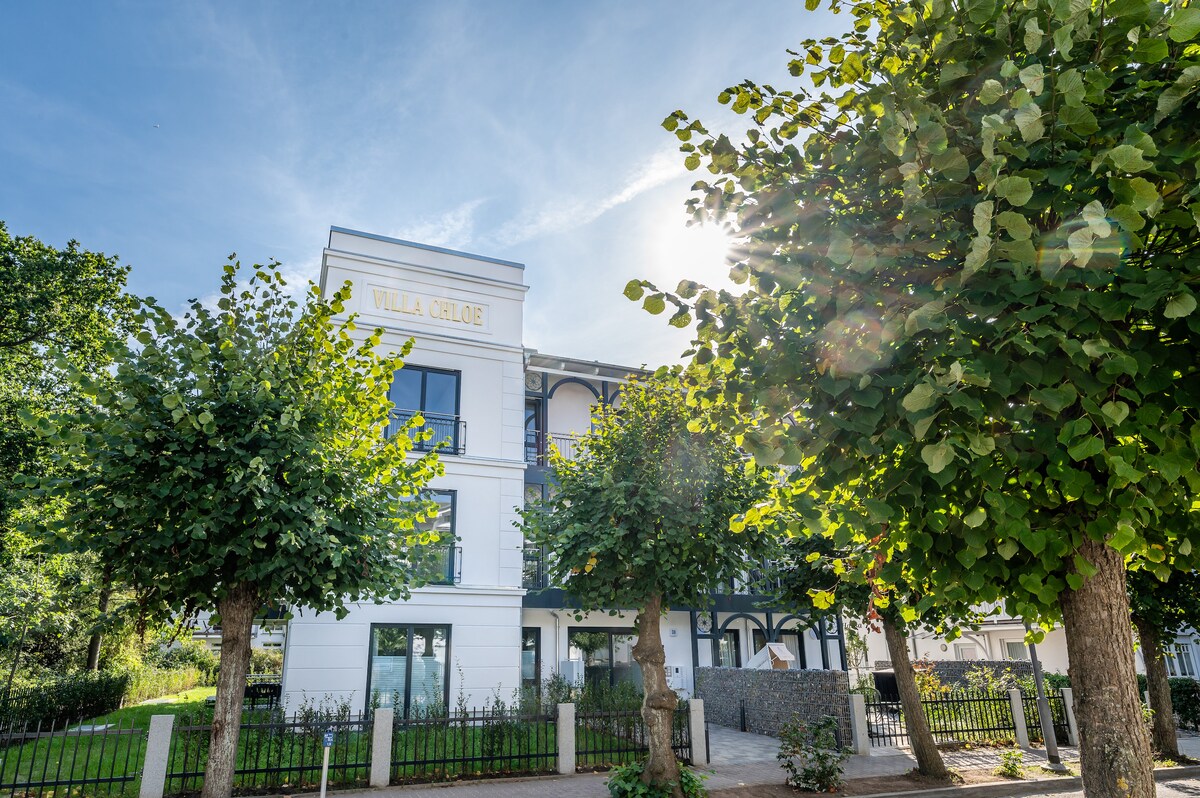The exterior of Villa Chloe is shown, featuring a modern architectural design with large windows. Lush green trees frame the building, which is illuminated by sunlight. A serene setting is highlighted, with well-maintained landscaping and a barrier-free entrance.