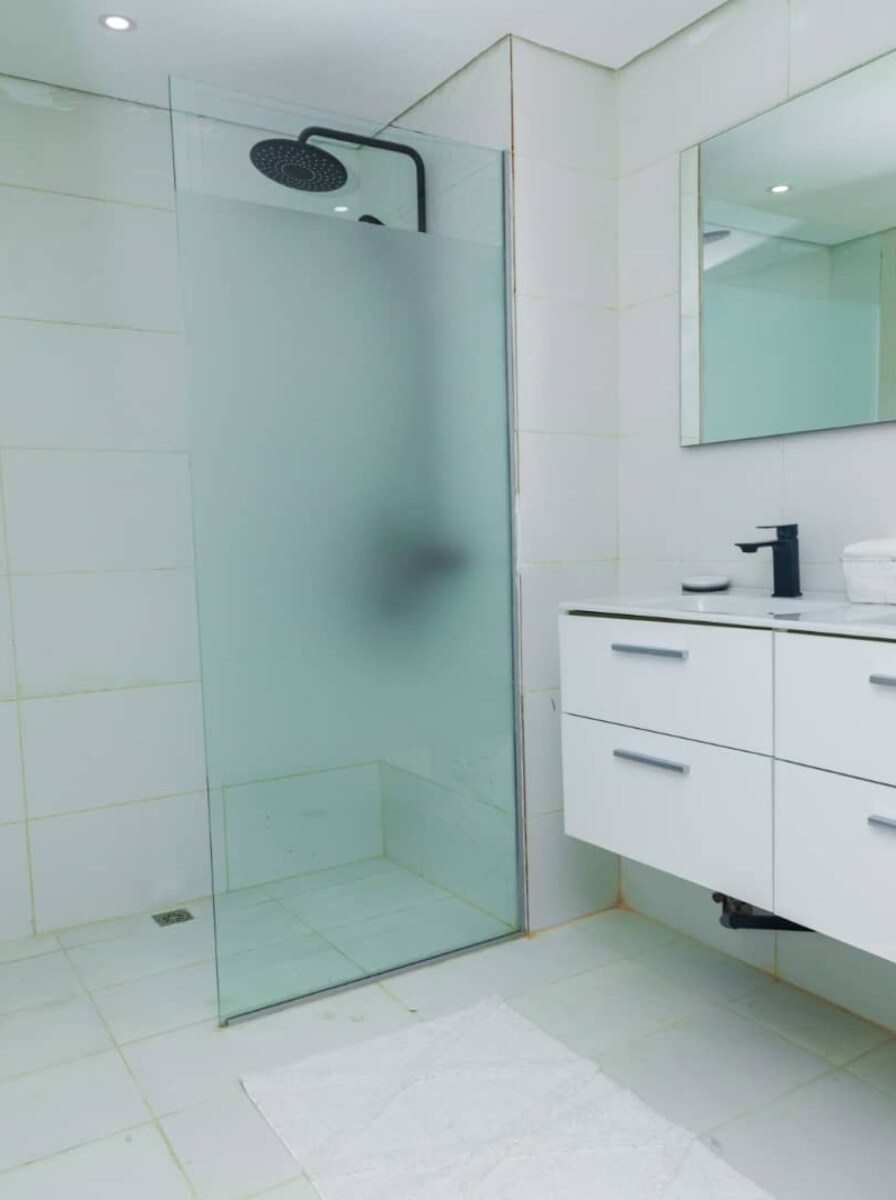 A modern bathroom is showcased with a glass shower enclosure featuring a rainfall showerhead. A sleek vanity with clean lines holds a black faucet and a framed mirror, while white tiles create a bright and airy atmosphere. Soft towels are neatly arranged nearby.