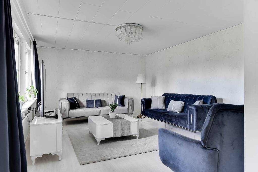 A bright and airy living room features a combination of plush blue sofas and a matching armchair, arranged around a soft area rug. A modern white coffee table sits at the center, and natural light filters through large windows, complemented by light-colored walls.