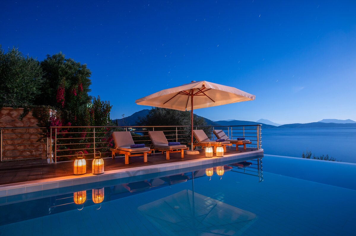 The infinity pool reflects the night sky, surrounded by lounge chairs and lanterns that provide soft illumination. An umbrella offers shade over part of the seating area, while lush greenery and the distant mountains enhance the serene coastal view.