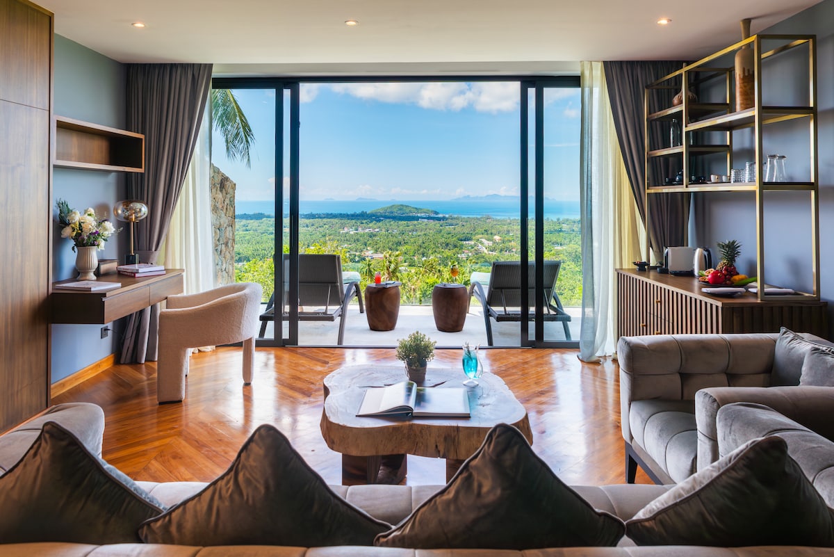 A spacious living area features a gray sectional sofa arranged around a light-toned coffee table. Large glass doors lead to a private terrace with views of lush greenery and distant sea. Natural light fills the room, complemented by elegant furnishings and decor.