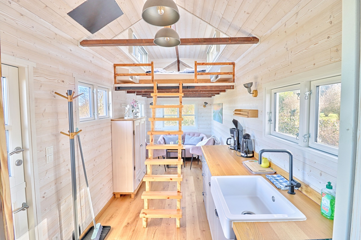An interior view of the tiny house features a wooden staircase leading to a sleeping loft. Natural light enters through multiple windows, illuminating a spacious kitchen area with a sink and countertop. A cozy seating area with comfortable couches is positioned nearby.
