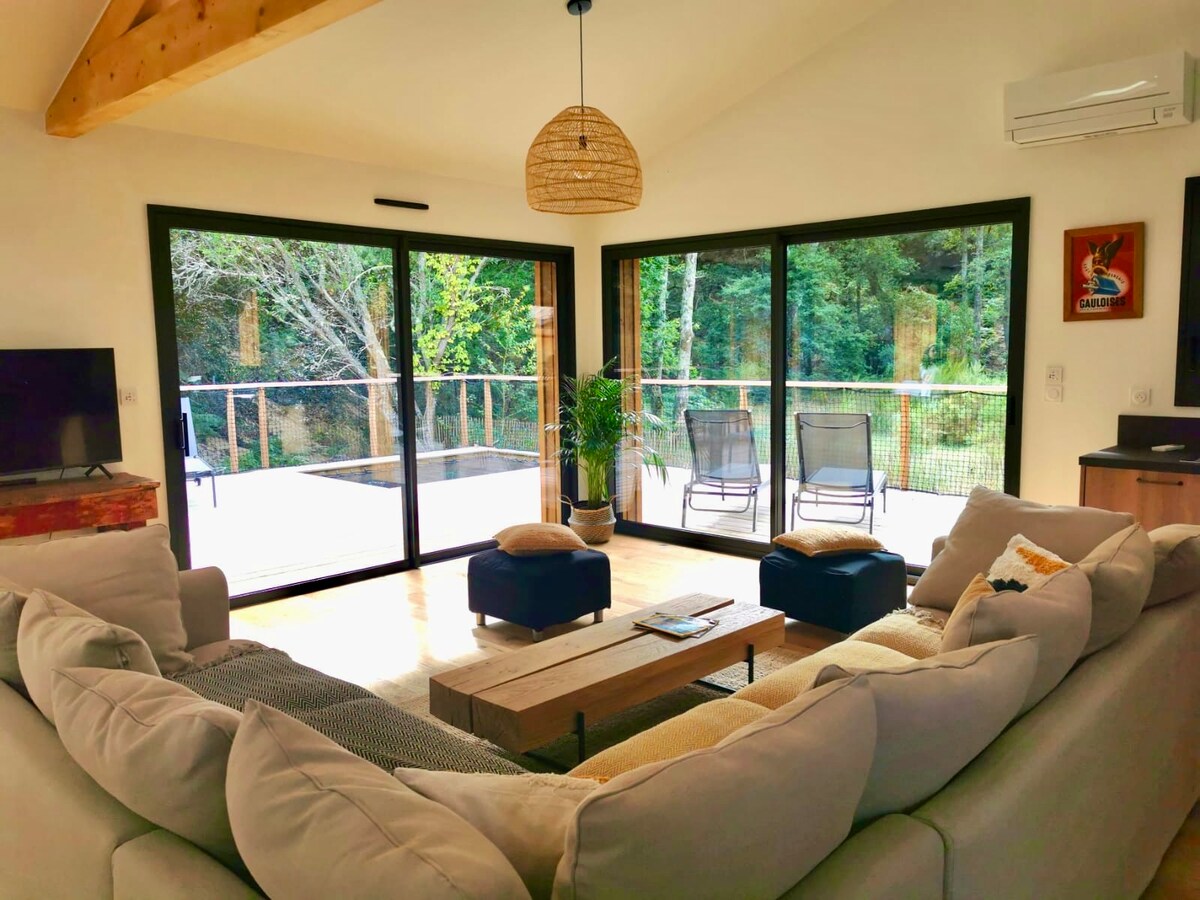 A spacious living area is showcased, featuring a large, light-coloured sectional sofa arranged in a circular layout. Floor-to-ceiling glass doors offer views of the surrounding greenery, while a wooden coffee table sits at the center. Two comfortable chairs are positioned nearby, and a subtle decorative lamp is visible.