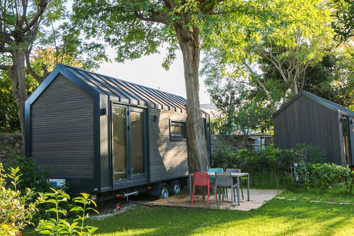 A compact tiny house is nestled among lush greenery. The exterior features dark wooden cladding and large windows. A small outdoor seating area is presented with colorful chairs and a table, surrounded by well-maintained plants and trees.