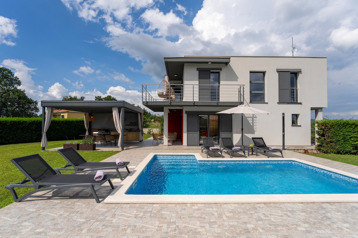 The exterior of a modern, two-story villa is showcased, featuring a private swimming pool bordered by lounge chairs. A covered outdoor dining area is seen on the left, and the lush green lawn surrounds the pool, creating a serene setting under a partly cloudy sky.