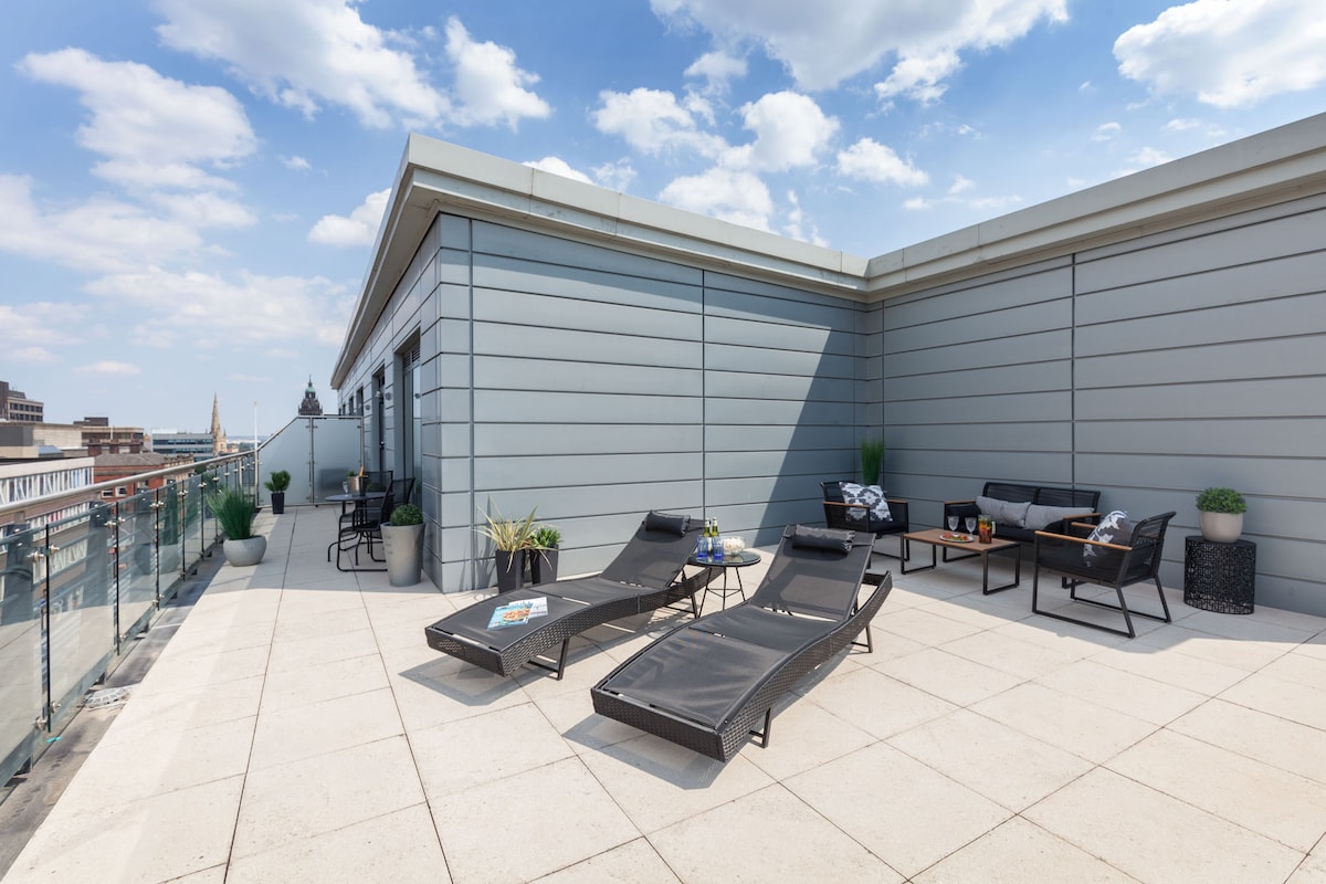 An expansive terrace features modern lounge chairs and a cozy seating area, surrounded by planters. The railing offers views of the city skyline under a partly cloudy sky, creating a serene outdoor space for relaxation.