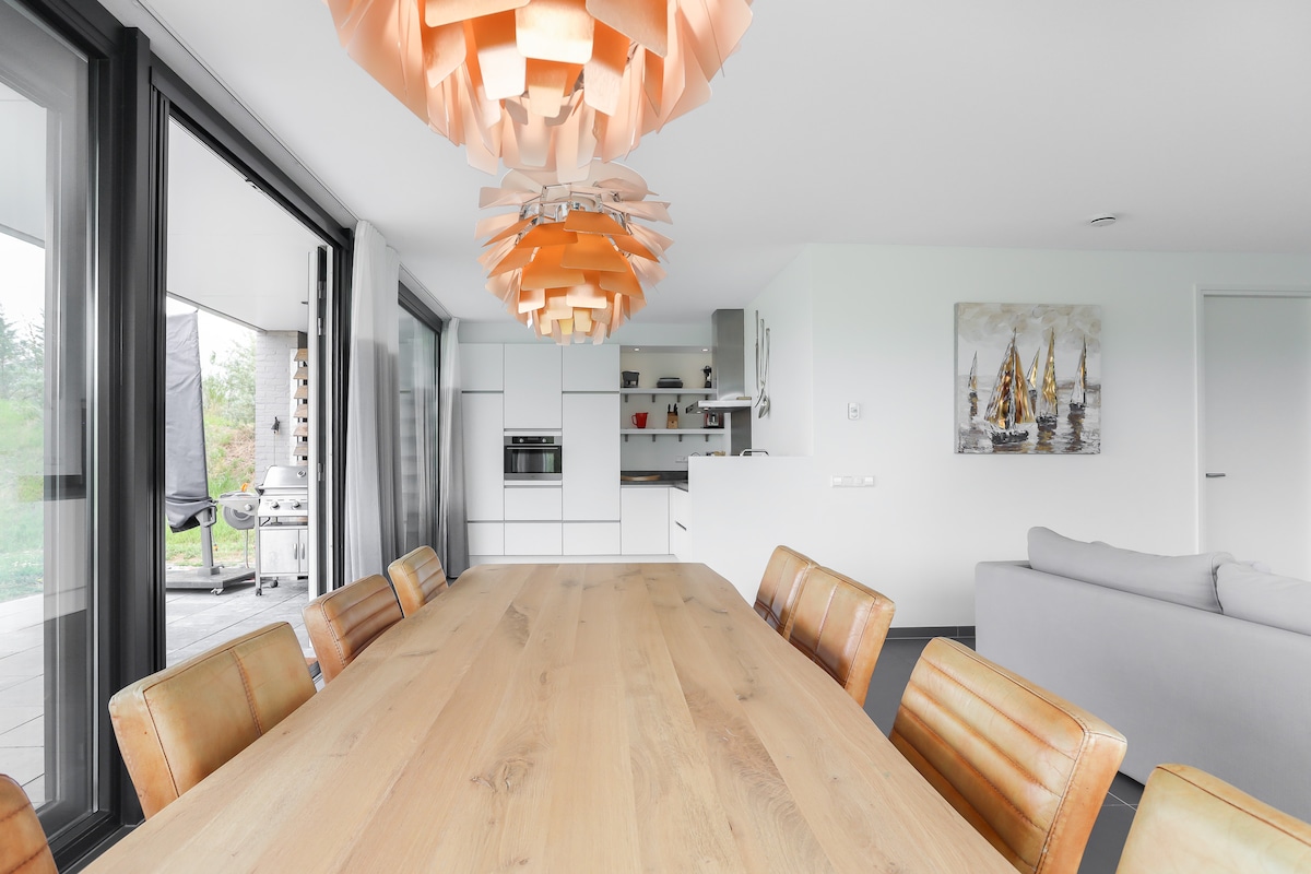 A spacious dining area is highlighted by a large wooden table surrounded by leather chairs. A modern kitchen is visible in the background, featuring sleek cabinetry and built-in appliances. Large glass doors provide a view of the outdoor space, enhancing the light-filled atmosphere.
