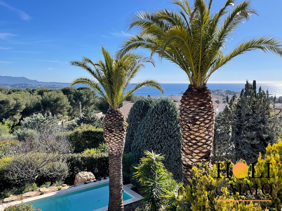Two tall palm trees are framed against a clear blue sky, overlooking a scenic hillside. The view reveals a glistening ocean in the distance, surrounded by lush greenery and vibrant foliage. A tranquil swimming pool is visible in the foreground, nestled within the garden.