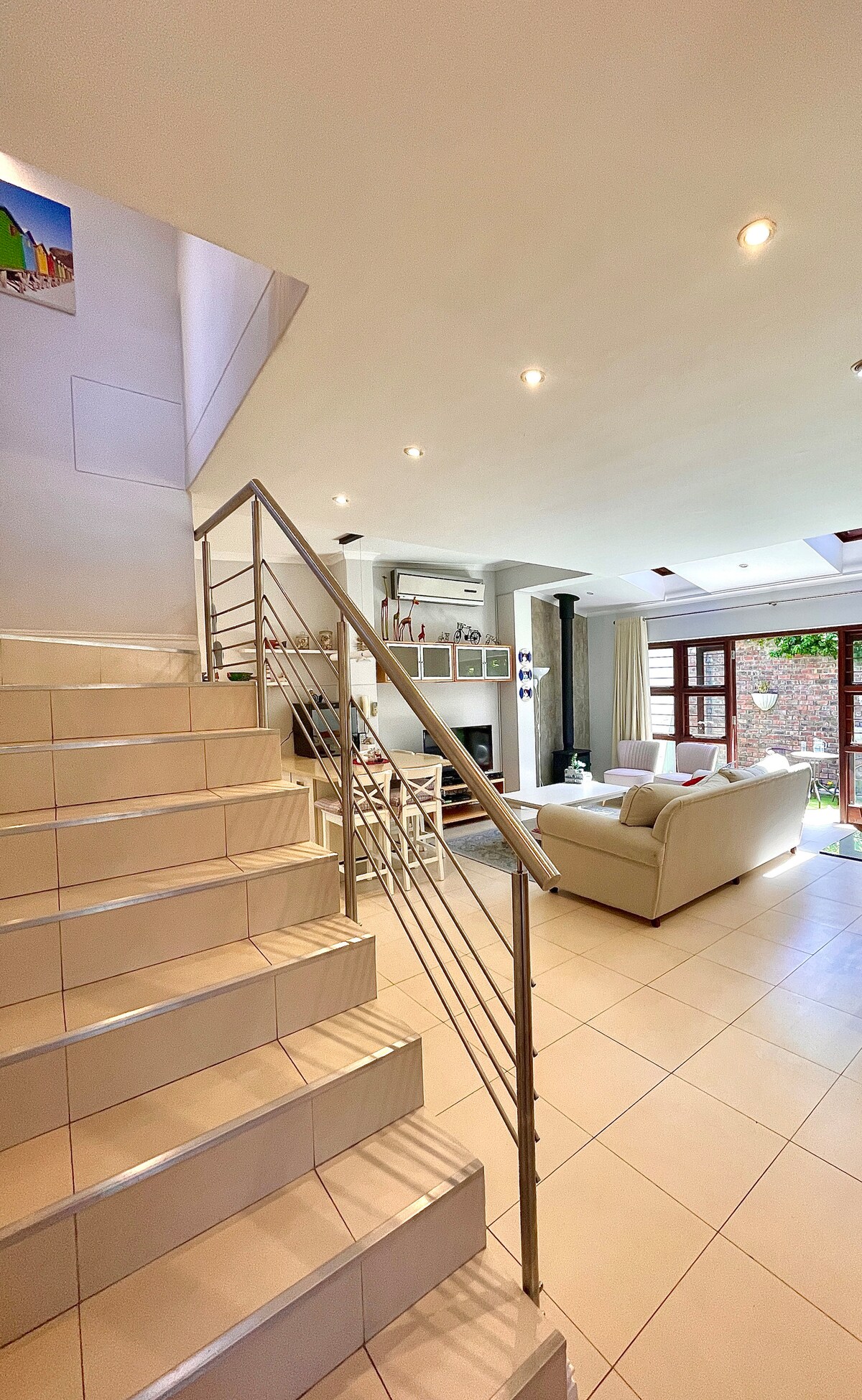 A well-lit stairway leads to a spacious lounge area, featuring a comfortable seating arrangement. Natural light floods the room through large garden-access windows and skylights, enhancing the open feel. The area is complemented by neutral-toned furnishings and contemporary decor.