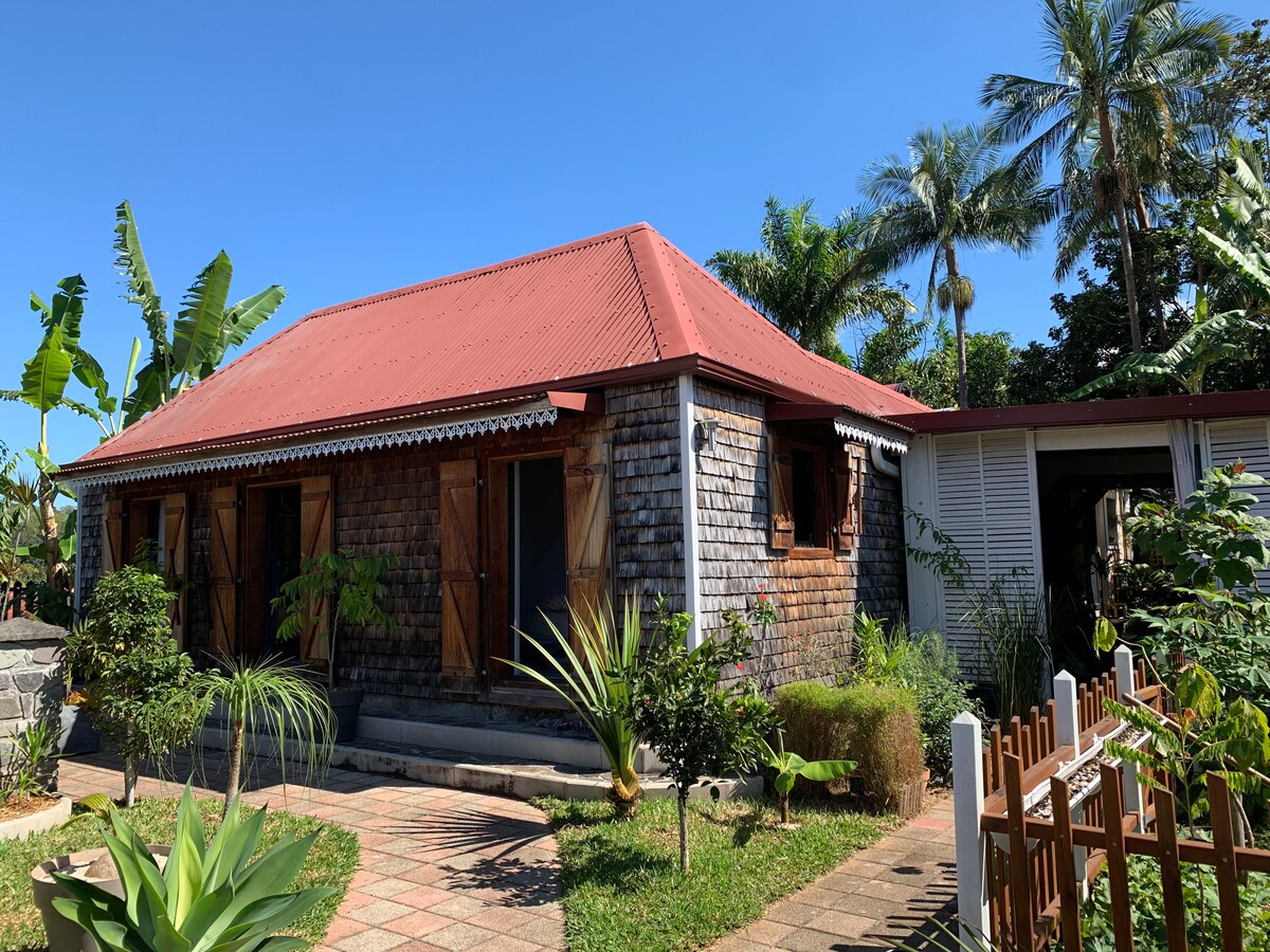 A charming exterior view of Ti Kaz Ananas is presented, showcasing its wooden facade with a vibrant red roof. Tropical plants enhance the landscape, while shuttered windows provide a rustic touch. The inviting pathway leads to the entrance, surrounded by lush greenery.