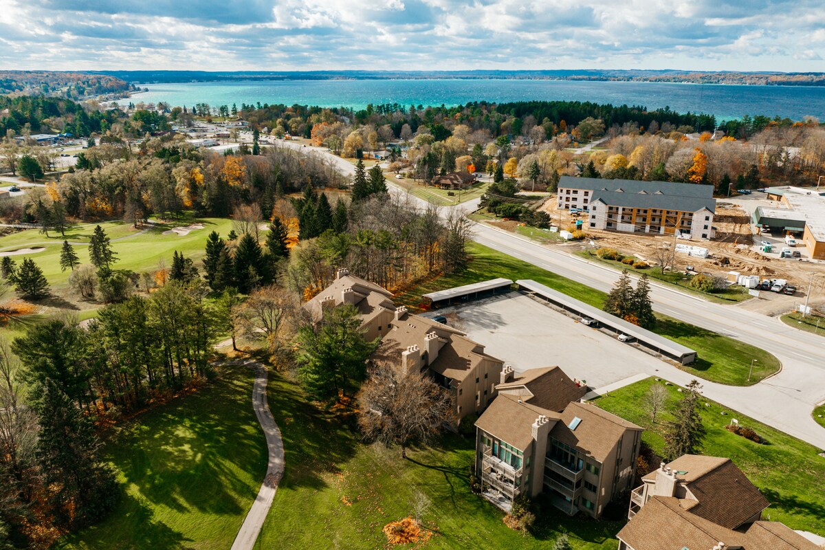 An aerial view captures the Golfside Getaway surrounded by green golf course areas and trees. The scenic Lake Michigan glimmers in the background, with a blend of autumn foliage. Nearby buildings and a road are also visible, enhancing the tranquil setting.