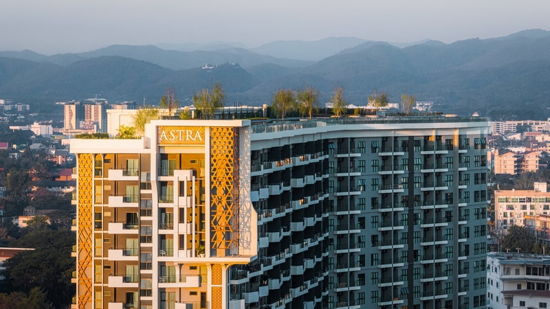 An aerial view displays a modern condominium building featuring a blend of architectural styles. Lush greenery crowns the rooftop, while the surrounding landscape includes distant mountains and urban structures, reflecting the vibrant environment of Chiang Mai.