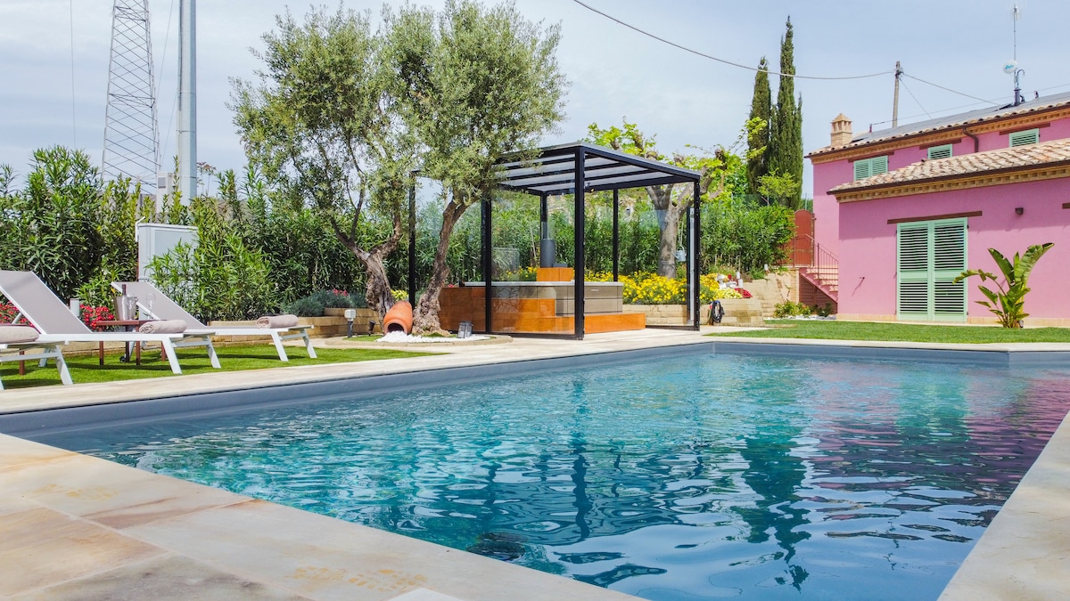 A clear pool reflects the surrounding greenery, with comfortable lounge chairs positioned nearby. A modern gazebo sits beside the pool, providing shade, while vibrant flowers and plants enhance the landscape. A charming pink building can be seen in the background.