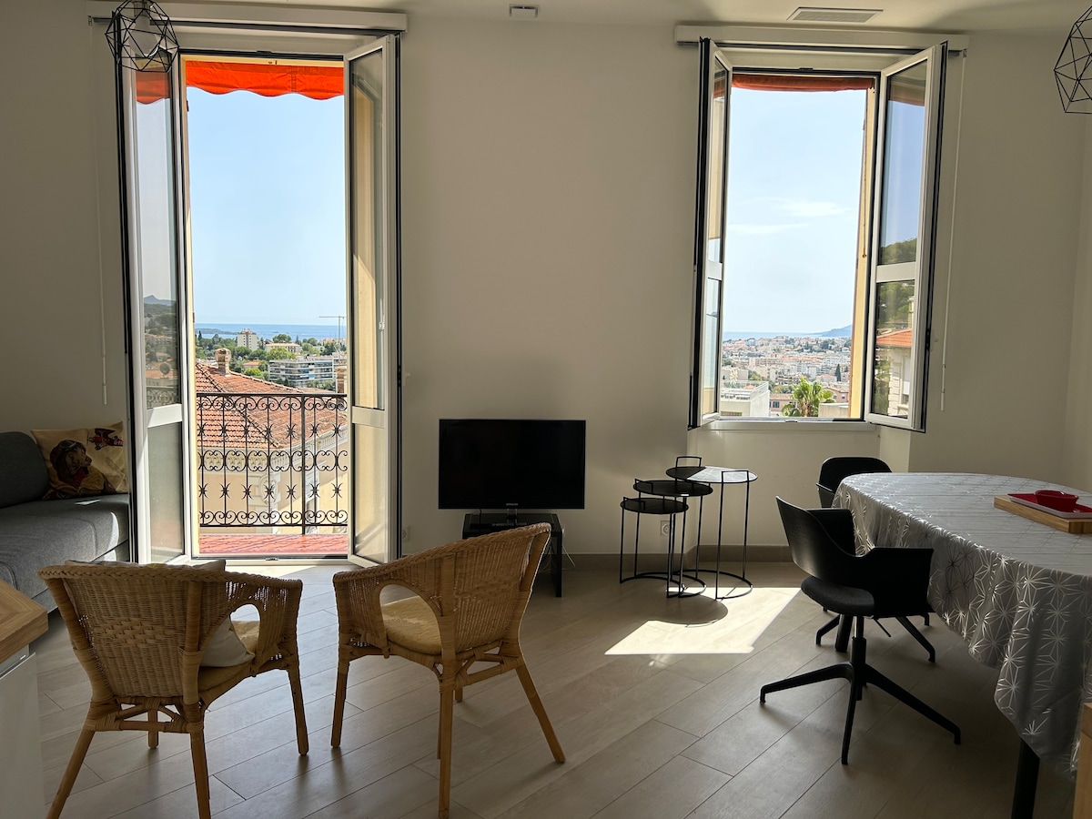 A bright living area is presented with two large windows offering scenic views of Cannes. A compact television is positioned near a table and chairs, while additional seating is provided by two rattan chairs. Natural light fills the space, creating a welcoming ambiance.