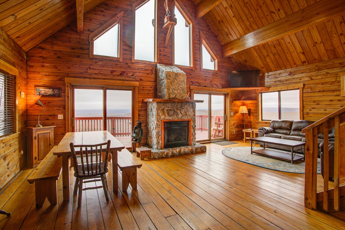 A spacious living area features high wooden ceilings and large windows that offer scenic views. A stone fireplace serves as a focal point, accompanied by a comfortable seating arrangement. A large dining table and wooden chairs are positioned near the windows, enhancing the cabin's cozy atmosphere.