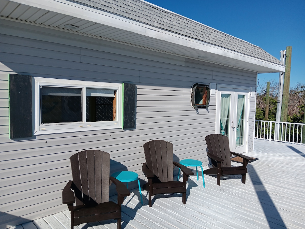 A spacious deck features two dark wooden Adirondack chairs paired with light blue side tables, offering a comfortable outdoor seating area. A window with shutters is positioned near the chairs, enhancing the outdoor ambiance while allowing natural light to enter the interior.