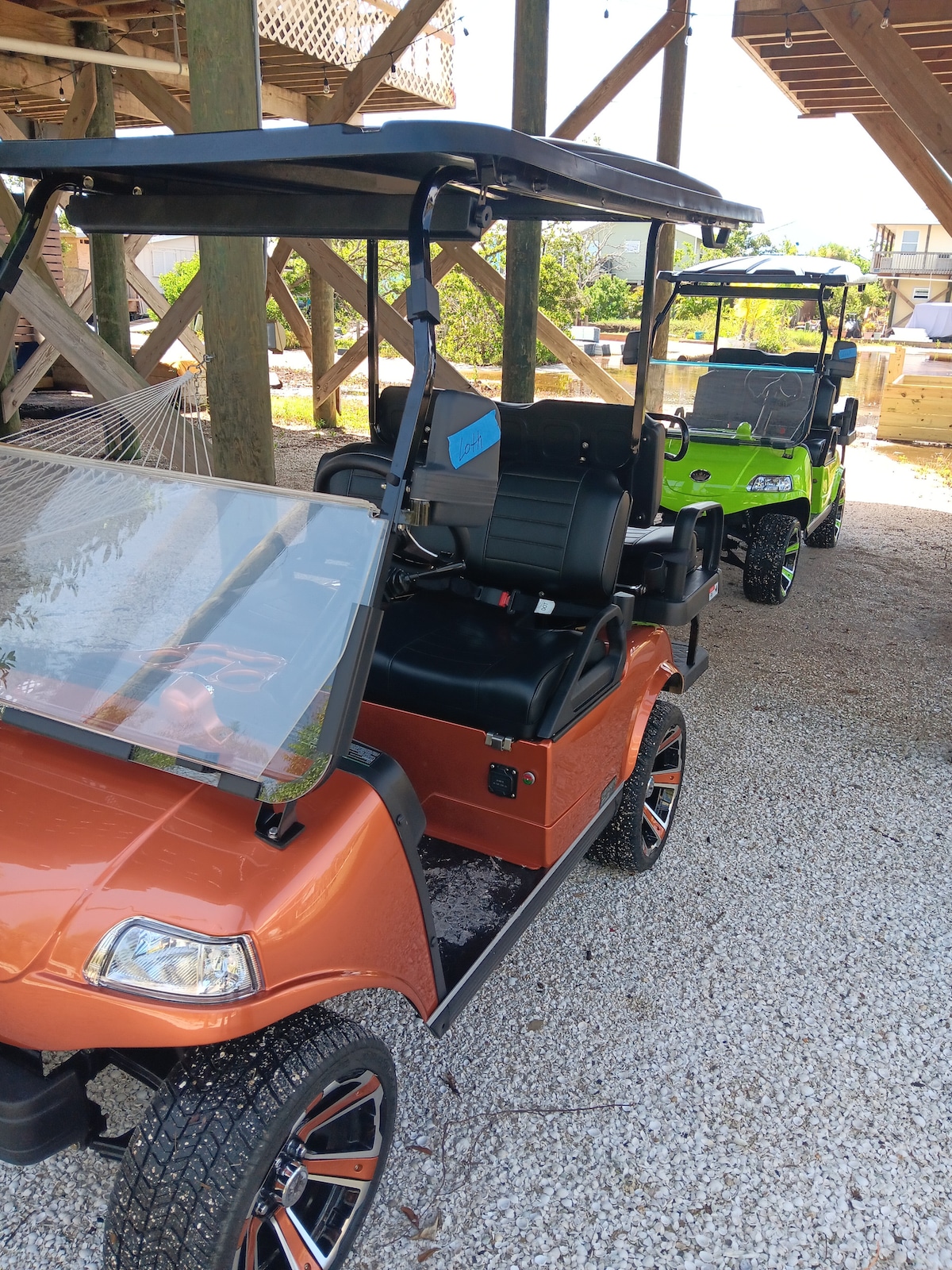 Two golf carts are parked on gravel beneath a raised structure, one in vibrant orange and the other in bright green. The carts feature sleek designs and comfortable seating, ready for exploration on the island.