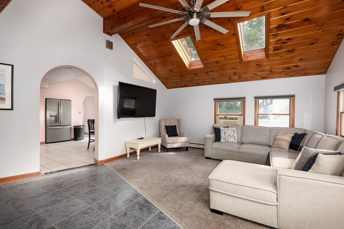 A cozy living room features a large sectional sofa and high ceilings with wooden beams. Natural light enters through multiple windows, while a flat-screen TV is mounted on the wall. An archway leads into the kitchen area, visible in the background.