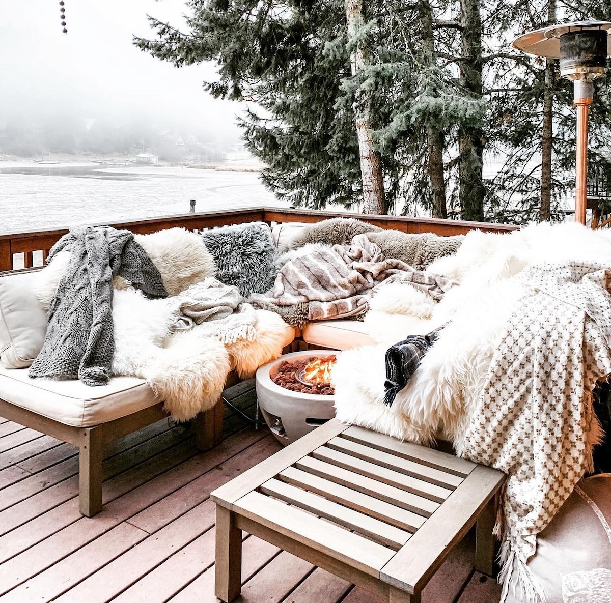 A cozy outdoor seating area is surrounded by tall trees, featuring soft blankets draped over plush cushions. A circular fire pit sits at the center, providing warmth, while the tranquil lake view is partially shrouded by mist in the background.