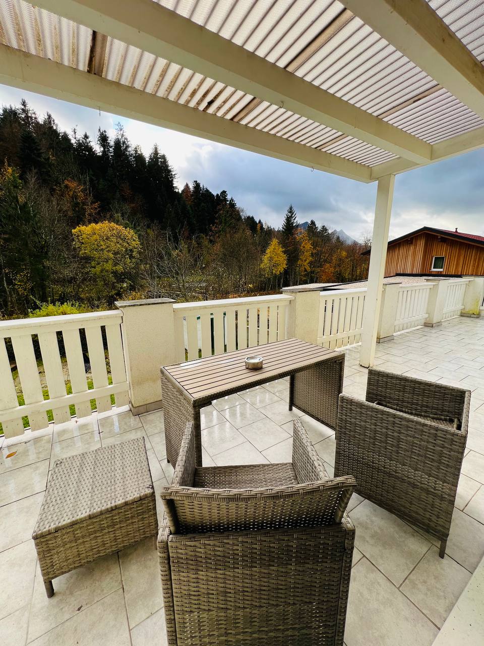 A spacious terrace features a small table set surrounded by four wicker chairs. The area is covered by a slatted roof, providing shade and a view of the surrounding trees and hills in the distance.