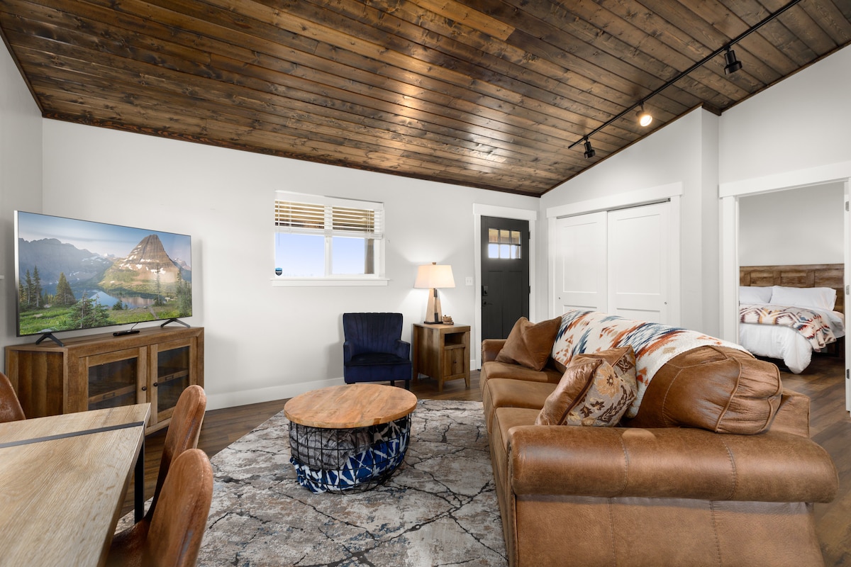 A spacious living area features a leather sofa and a round coffee table, with a dark blue accent chair nearby. A television displaying mountain scenery is mounted on the wall. Natural light streams in through a window, and a door leads to an adjoining room.