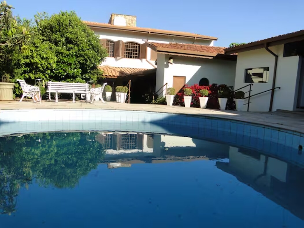A serene outdoor area features a clear swimming pool reflecting the surrounding greenery and a well-maintained garden. Comfortable seating options are visible nearby, along with potted plants that add color to the space. The building's architecture complements the inviting environment.