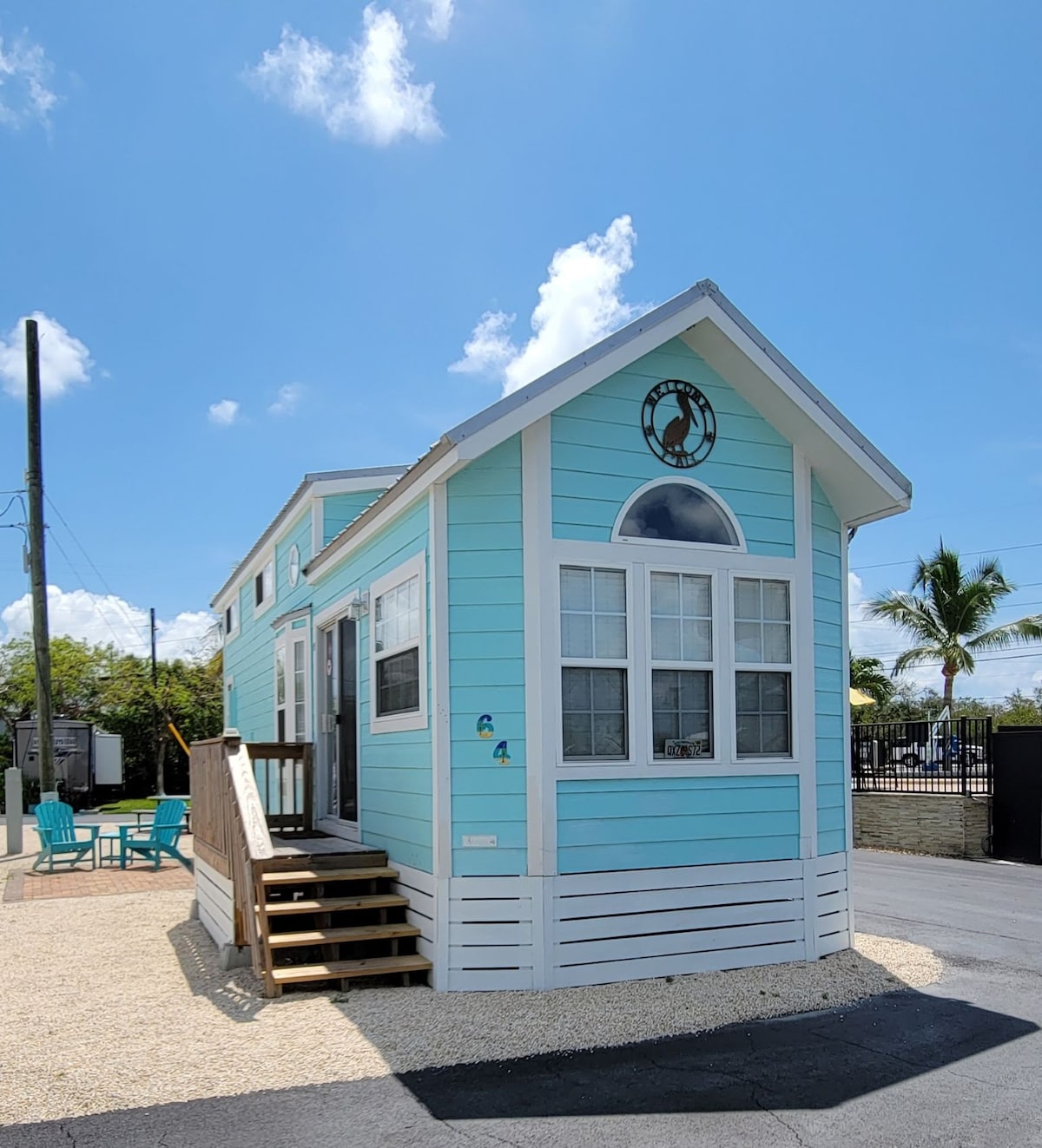 A charming exterior of a light blue house is shown against a clear sky. The house features large front windows, a decorative round window, and a small front porch with steps leading up. Outdoor seating in turquoise is visible nearby, set on a gravel driveway.