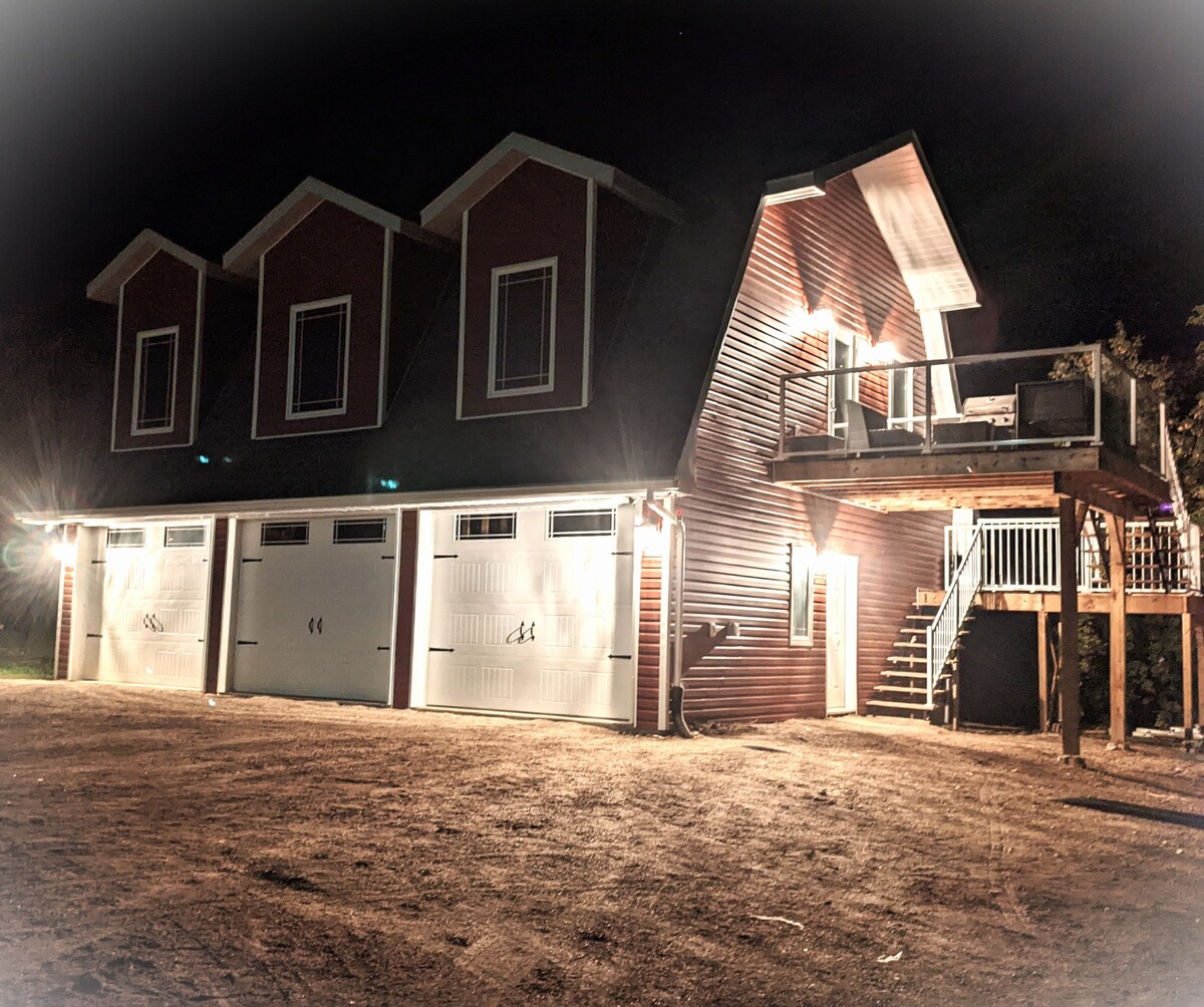 The exterior of a multi-level lodge is illuminated at night, featuring three garages with white doors. A wooden deck is visible above the garage, accessed by a staircase. Soft lighting outlines the structure, set in a rural landscape.