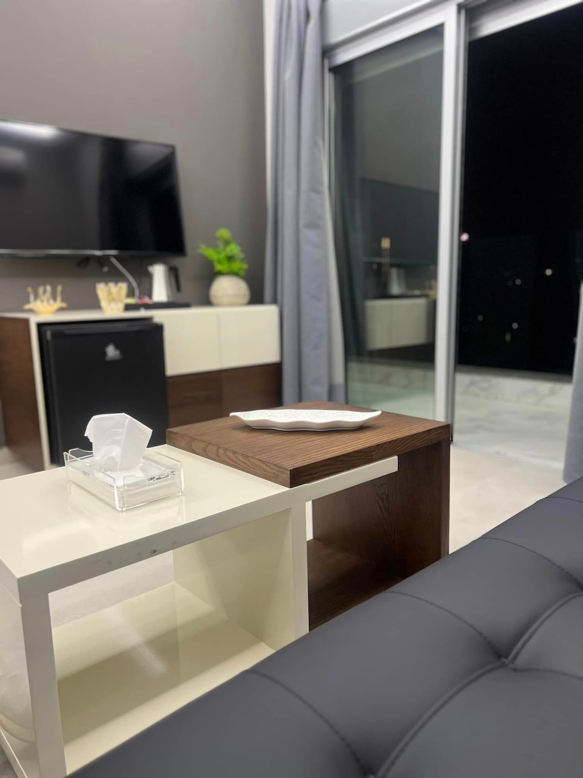 A modern living area is showcased with a sleek black sofa and a minimalist coffee table. A flat-screen TV is mounted on the wall, while a small refrigerator is visible in the background. Natural light streams in through a partially open door leading to a balcony.