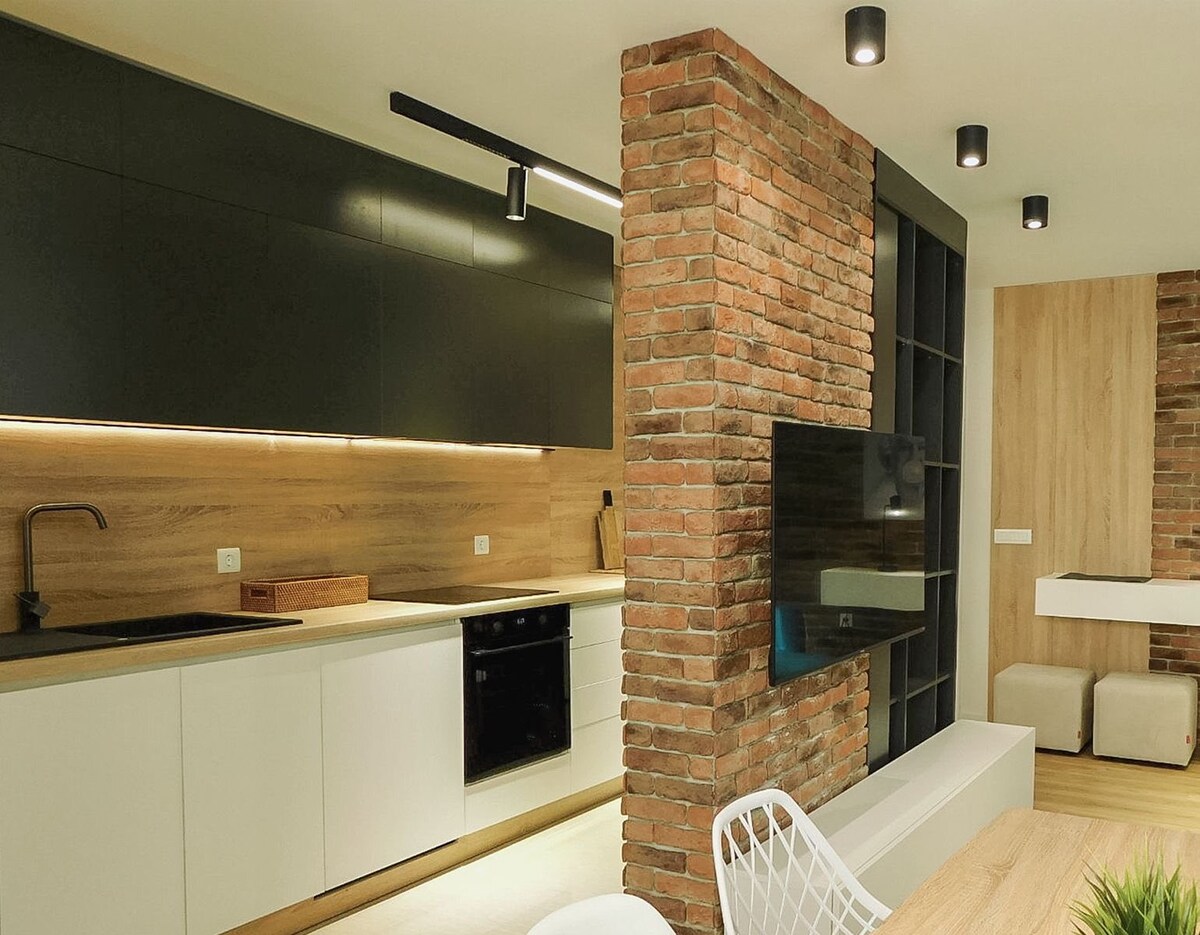 A modern kitchen area is highlighted by sleek black cabinets and a contrasting brick accent wall. Under-cabinet lighting illuminates the countertop space, while a flat-screen television is mounted on the adjacent wall. A wooden dining table with white chairs is positioned in the foreground.