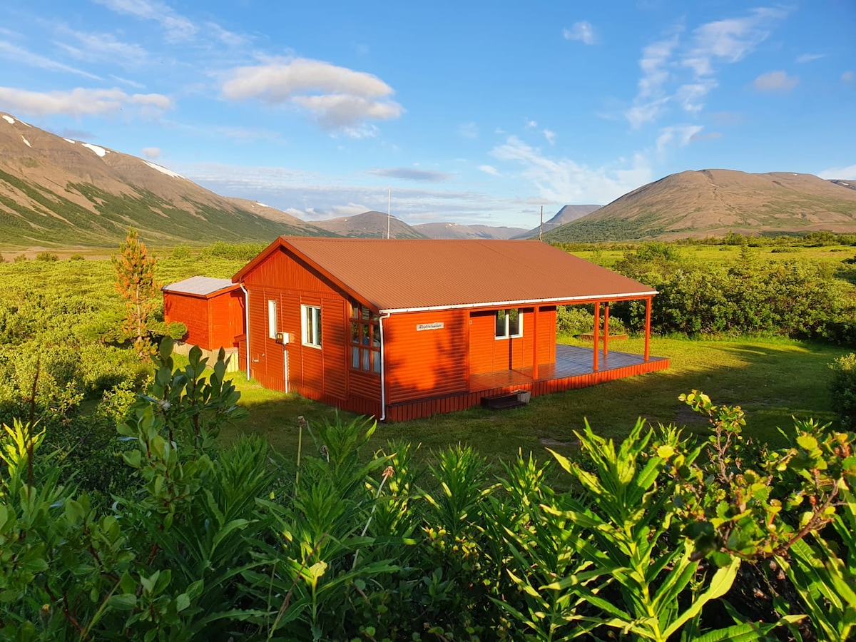 A charming summer house, painted in vibrant red, is surrounded by lush greenery and rolling hills. The structure features a covered porch at the front, with scenic mountain views extending into the distance. The tranquil setting offers a peaceful escape into nature.