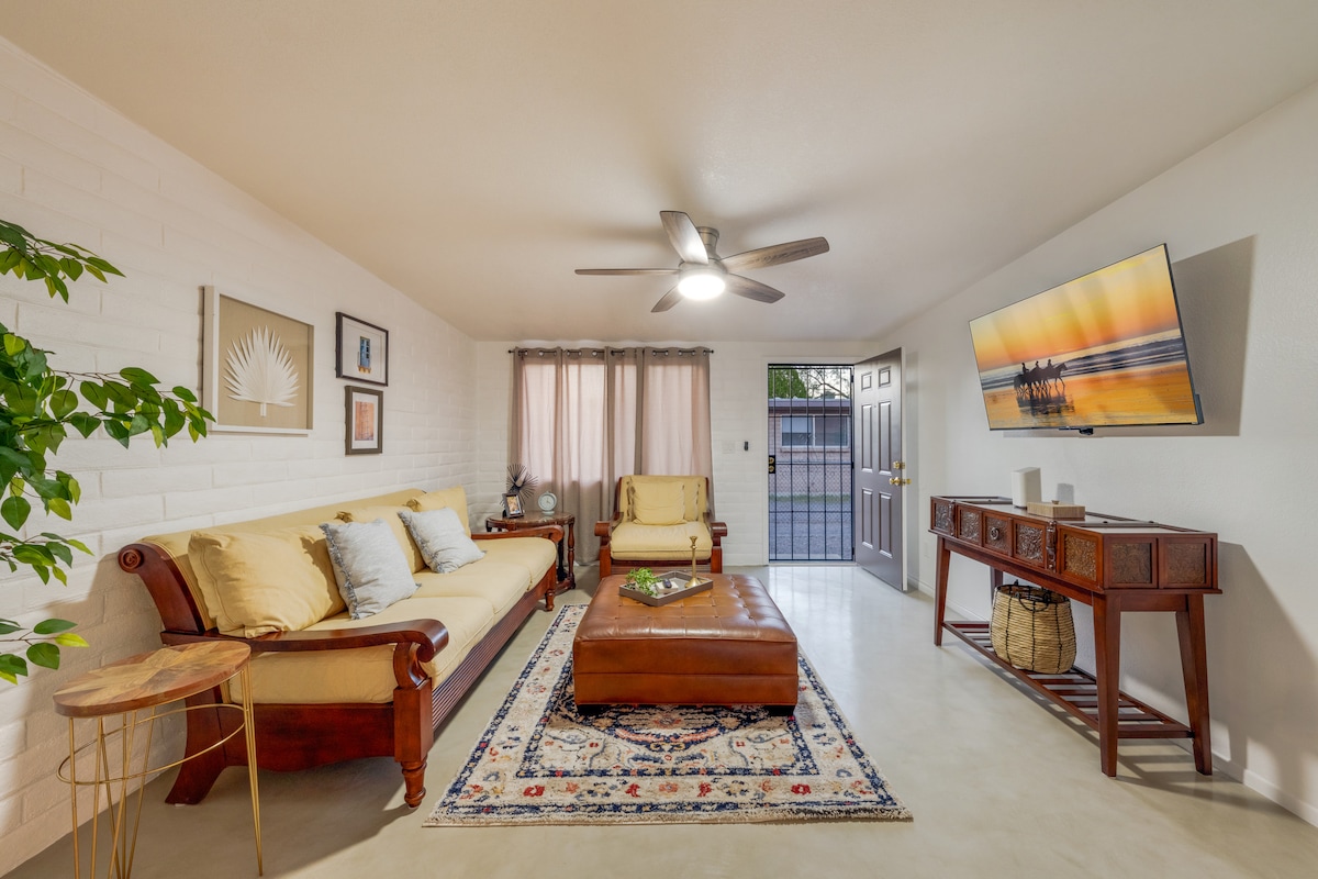 A bright living area features a comfortable sofa and a stylish leather coffee table. A large smart TV is mounted on the wall. Natural light filters through sheer curtains, complementing the warm tones of the decor and the textured white walls.