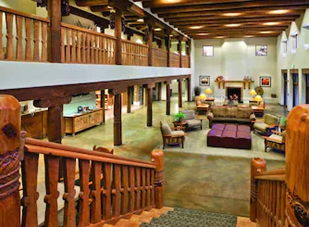 A spacious living area is illuminated by natural light from high windows, featuring a central seating arrangement with a large, cozy sofa and armchairs. Exposed wooden beams accent the ceiling, and a fireplace is positioned against the back wall, enhancing the communal atmosphere.